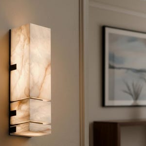 Op de afbeelding: Een moderne wandlamp met een rechthoekig, gelaagd ontwerp. De lamp is gemaakt van een doorschijnend, gebroken wit materiaal met bruine adering, gemonteerd op een zwart metalen frame. De lamp is verlicht en geeft een warme gloed.