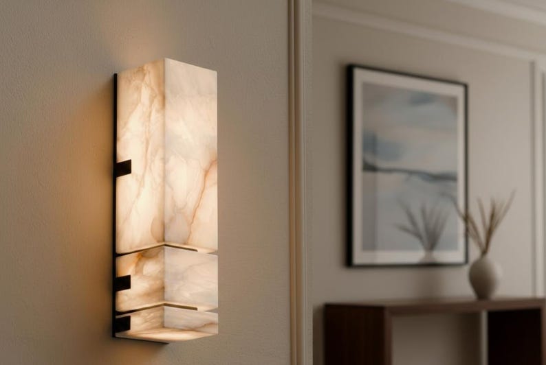 Op de afbeelding: Een moderne wandlamp met een rechthoekig, gelaagd ontwerp. De lamp is gemaakt van een doorschijnend, gebroken wit materiaal met bruine adering, gemonteerd op een zwart metalen frame. De lamp is verlicht en geeft een warme gloed.