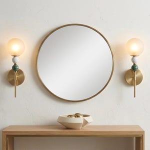 May include: A round, gold-framed mirror hangs above a wooden console table. Two brass sconces with frosted glass globes and decorative stone accents flank the mirror. A geometric bowl with gold spheres sits on the table.