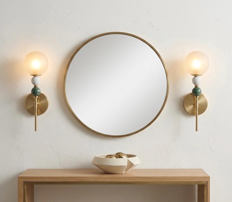 May include: A round, gold-framed mirror hangs above a wooden console table. Two brass sconces with frosted glass globes and decorative stone accents flank the mirror. A geometric bowl with gold spheres sits on the table.