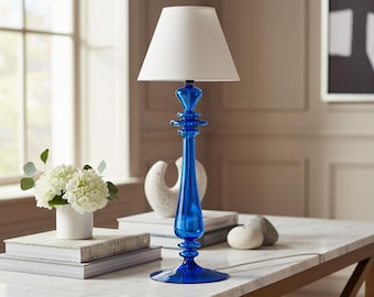 May include: A blue glass table lamp with a white lampshade. The lamp has a decorative, ornate base. The lamp is on a marble-topped table with books, a vase of flowers, and decorative objects. The room has a window and artwork.
