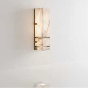 Op de afbeelding: Een rechthoekige wandlamp met een lichtgekleurde, doorschijnende kap. De lamp heeft een messingkleurig metalen frame en bevestigingsmateriaal. De lamp is aan een witte muur bevestigd. De lamp is ongeveer 61 cm hoog.