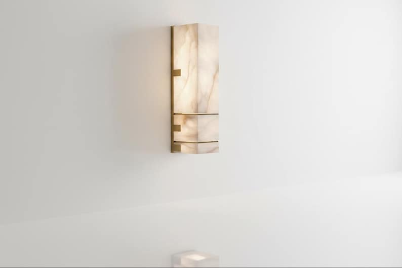 Op de afbeelding: Een rechthoekige wandlamp met een lichtgekleurde, doorschijnende kap. De lamp heeft een messingkleurig metalen frame en bevestigingsmateriaal. De lamp is aan een witte muur bevestigd. De lamp is ongeveer 61 cm hoog.