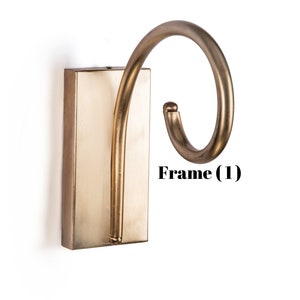 May include: A wall-mounted, antique brass-colored hook with a rectangular base and a curved, swirling hook design. The hook is mounted on a white background. The text "Frame (1)" is visible.