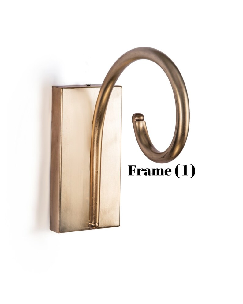 May include: A wall-mounted, antique brass-colored hook with a rectangular base and a curved, swirling hook design. The hook is mounted on a white background. The text "Frame (1)" is visible.