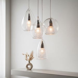 May include: Four clear glass pendant lights with chrome accents hang from the ceiling. Each light has a unique shape, including teardrop, tulip, and spherical designs. The lights are illuminated, showcasing a textured interior. A small vase with dried flowers sits on a shelf below.