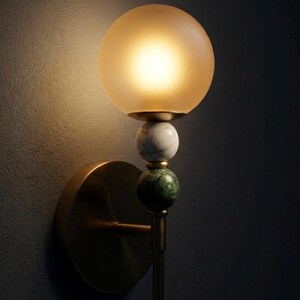 May include: A wall-mounted light fixture with a round, frosted glass shade emitting a warm glow. The fixture features a brass base and stem, with decorative marble and green stone spheres. The design is elegant and suitable for interior decor.