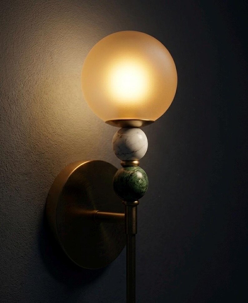 May include: A wall-mounted light fixture with a round, frosted glass shade emitting a warm glow. The fixture features a brass base and stem, with decorative marble and green stone spheres. The design is elegant and suitable for interior decor.