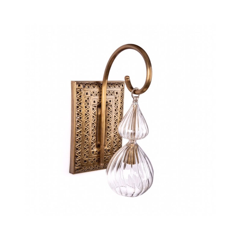 May include: A brass wall sconce with a rectangular backplate featuring a geometric pattern. A curved brass arm supports a clear, ribbed glass pendant with two bulbous sections. The sconce is designed for indoor lighting.