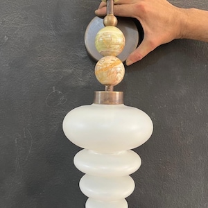 May include: A decorative wall sconce with a stacked design. The top has three spherical, marbled stone elements in green and tan, connected by a bronze rod. Below, a white, frosted glass shade with a layered, rounded shape.
