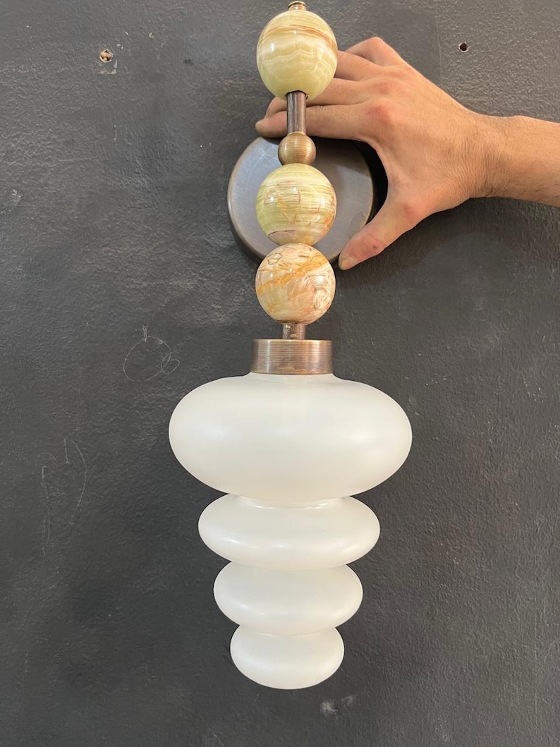 May include: A decorative wall sconce with a stacked design. The top has three spherical, marbled stone elements in green and tan, connected by a bronze rod. Below, a white, frosted glass shade with a layered, rounded shape.