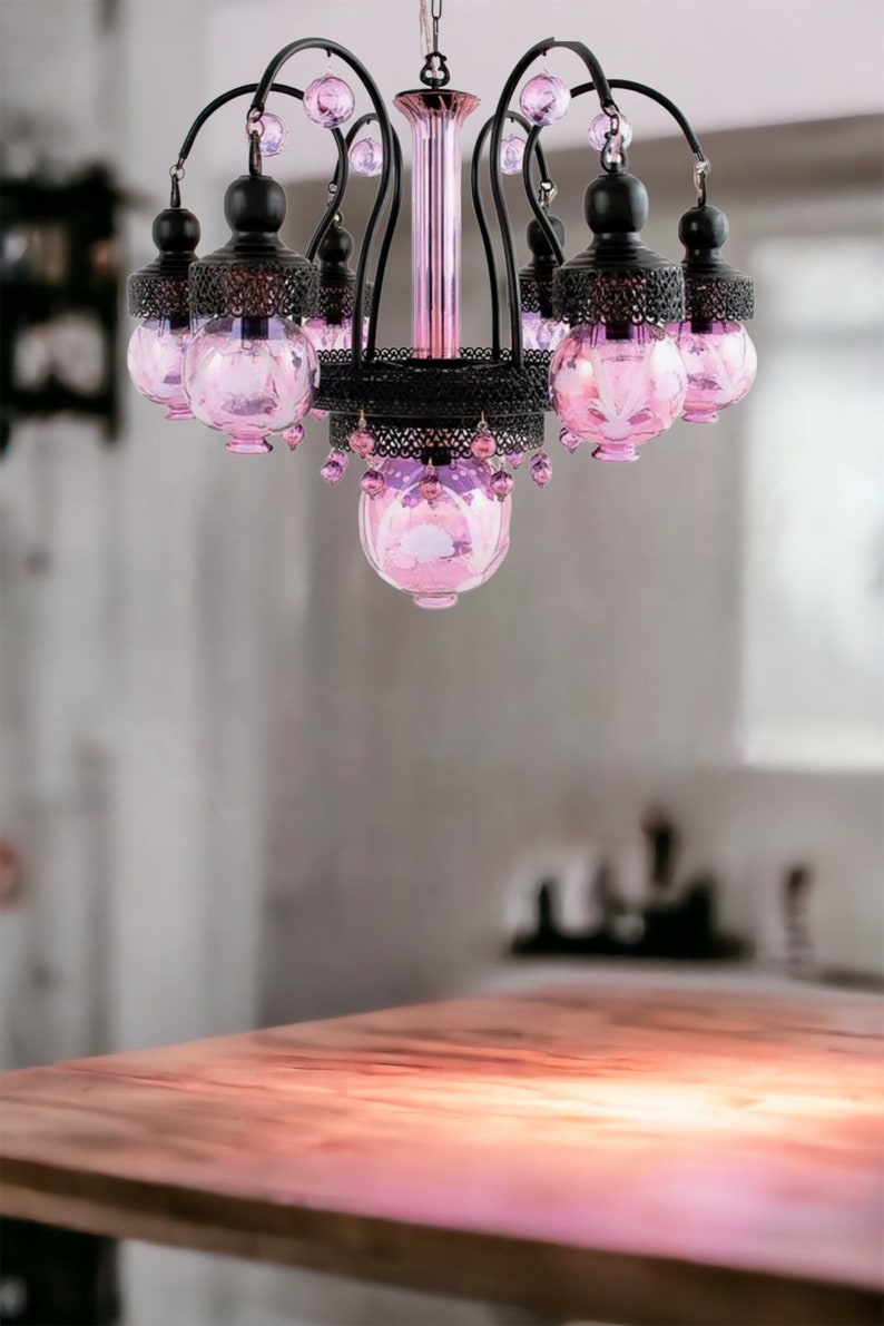 May include: Ornate chandelier with a black metal frame and pink glass shades. The chandelier has a central column and curved arms, each holding a pink glass globe. Decorative crystals hang from the bottom.