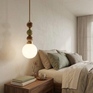 May include: A modern pendant light with a brass rod and decorative stone beads in shades of brown and green. The light fixture has a large, round, frosted glass bulb. The bedroom setting includes a wooden nightstand, books, and a bed with pillows.