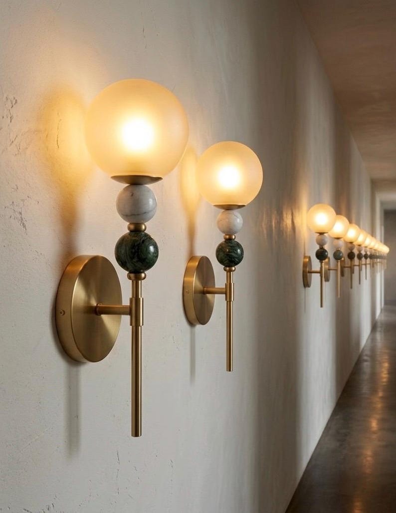 May include: A series of wall sconces with a modern design. Each sconce features a round, frosted glass globe, marble and green stone accents, and a brass finish. The sconces are mounted on a textured white wall, creating a warm, inviting ambiance.