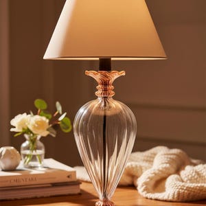 Könnte beinhalten: Eine dekorative Tischlampe mit einem transparenten, gerippten Glassockel und einem beigefarbenen Stoffschirm. Die Lampe hat einen roségoldfarbenen Akzent an der Basis und an der Oberseite. Die Lampe steht auf einer Holzoberfläche.