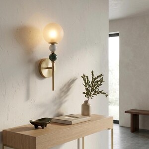 May include: A modern wall sconce with a frosted glass globe, brass stem, and decorative stone accents. The sconce is mounted on a textured white wall, providing ambient lighting. The design includes a brass base and a green and white stone detail.