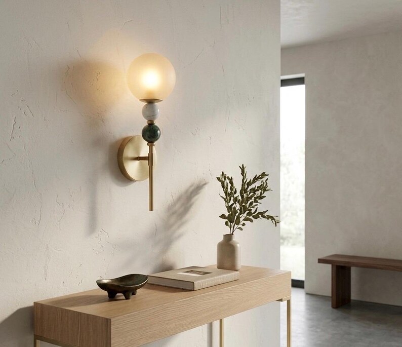 May include: A modern wall sconce with a frosted glass globe, brass stem, and decorative stone accents. The sconce is mounted on a textured white wall, providing ambient lighting. The design includes a brass base and a green and white stone detail.