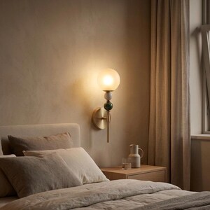 May include: A wall-mounted sconce with a frosted globe shade, brass fixture, and decorative green and white accents. The lamp is mounted on a beige wall above a bed with neutral-toned bedding.