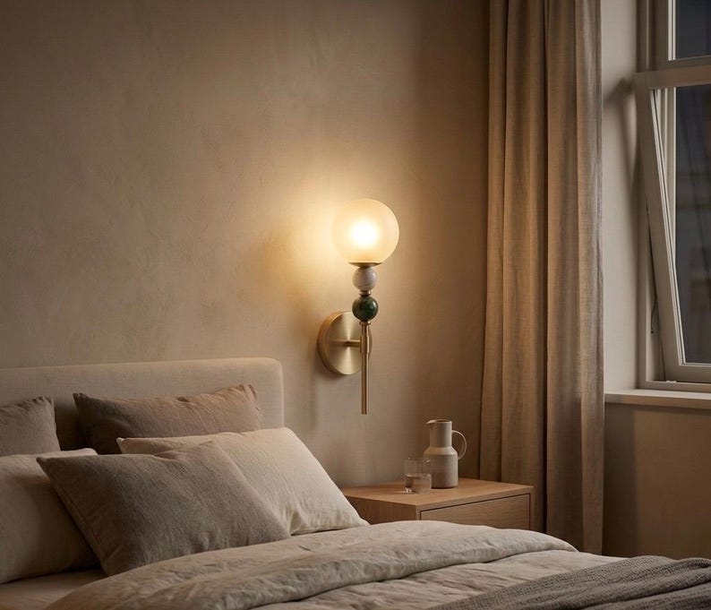 May include: A wall-mounted sconce with a frosted globe shade, brass fixture, and decorative green and white accents. The lamp is mounted on a beige wall above a bed with neutral-toned bedding.