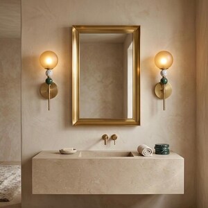 May include: A bathroom scene featuring a gold-framed mirror above a stone vanity. Two wall sconces with amber glass globes and decorative accents flank the mirror. The vanity has a built-in sink, with a rolled towel and decorative items.