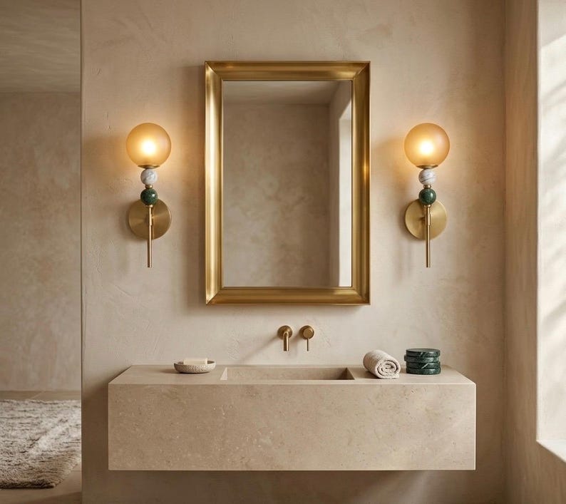 May include: A bathroom scene featuring a gold-framed mirror above a stone vanity. Two wall sconces with amber glass globes and decorative accents flank the mirror. The vanity has a built-in sink, with a rolled towel and decorative items.
