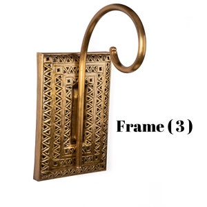 May include: A decorative wall sconce with a rectangular frame and a curved arm. The frame is a warm gold color with a geometric pattern. The text "Frame (3)" is visible on the right side of the image.