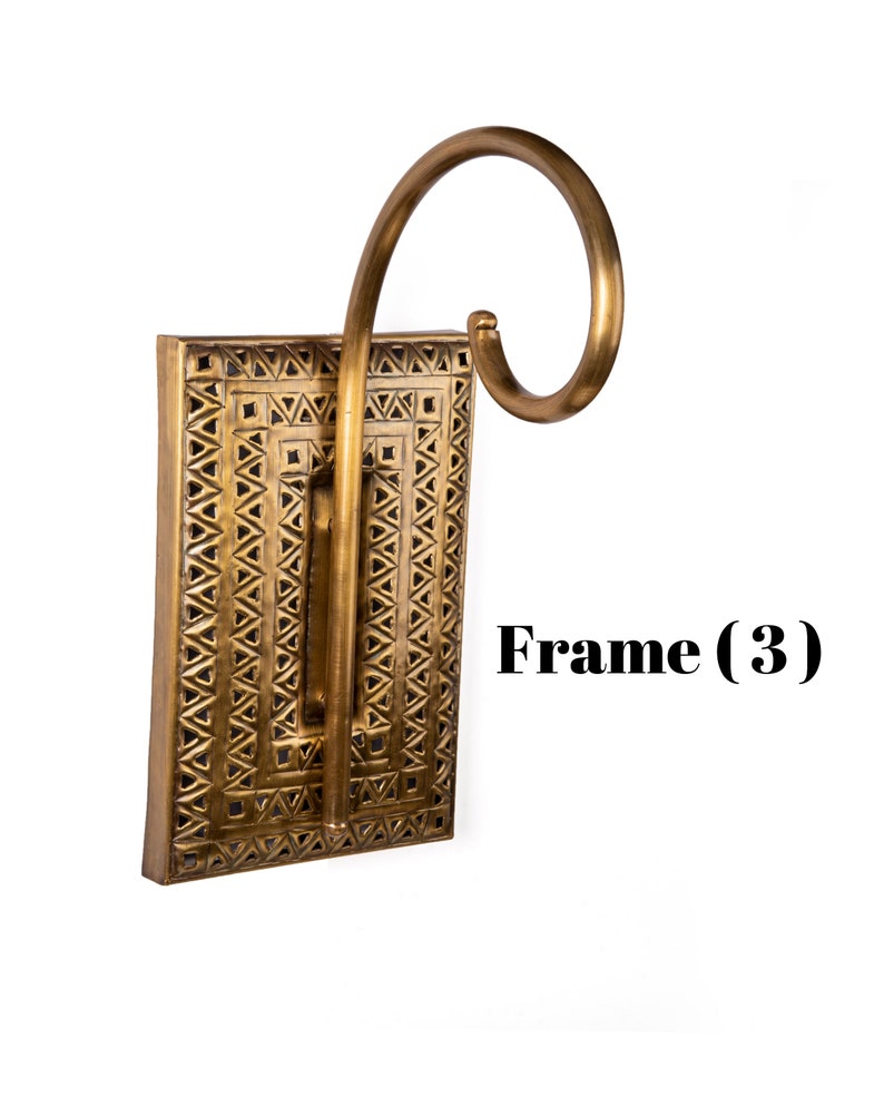 May include: A decorative wall sconce with a rectangular frame and a curved arm. The frame is a warm gold color with a geometric pattern. The text "Frame (3)" is visible on the right side of the image.