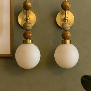 Könnte beinhalten: Zwei Wandleuchten im Mid-Century-Modern-Design. Jede Leuchte hat eine weiße Kugelleuchte, Messingakzente und dekorative Holzkugeln. Die Leuchten sind an einer grünen Wand montiert und schaffen eine warme Atmosphäre.