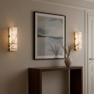 Op de afbeelding: Twee rechthoekige wandkandelaars met een marmerachtige uitstraling zijn aan een lichtgekleurde muur bevestigd. De wandkandelaars zijn verlicht en geven een warme gloed. Een ingelijste landschapsschilderij en een houten console met een vaas met gedroogde planten maken het interieur compleet.