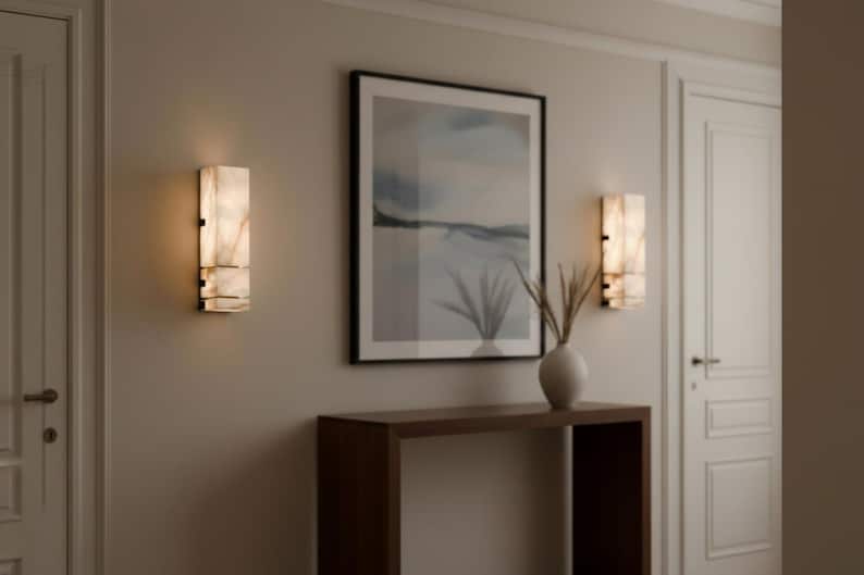 Op de afbeelding: Twee rechthoekige wandkandelaars met een marmerachtige uitstraling zijn aan een lichtgekleurde muur bevestigd. De wandkandelaars zijn verlicht en geven een warme gloed. Een ingelijste landschapsschilderij en een houten console met een vaas met gedroogde planten maken het interieur compleet.