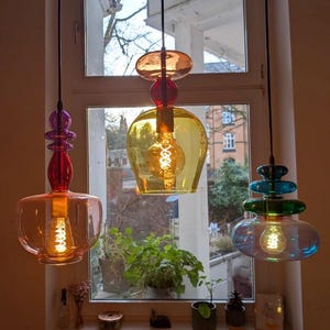 Puede incluir: Tres lámparas colgantes de cristal de colores cuelgan frente a una ventana. Las luces son en tonos naranja, amarillo y azul-verdoso, cada una con una forma única y una bombilla visible. La ventana da a un jardín.