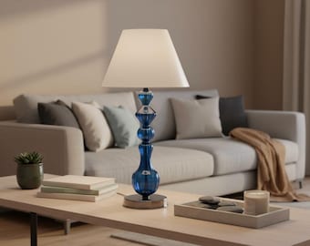 May include: A blue glass table lamp with a white lampshade sits on a light wood coffee table. The lamp has a decorative, stacked design. A sofa with pillows and a blanket is in the background.