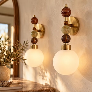 May include: Two wall sconces featuring a round brass mounting plate and a vertical stem adorned with three stacked decorative spheres in brown and green tones, topped with a large, spherical, frosted white glass light shade.