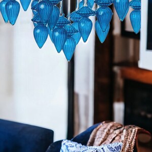 May include: A modern chandelier with multiple blue glass teardrop-shaped pendants. The pendants are suspended from a silver-toned metal frame. The chandelier hangs above a blue velvet chair with a patterned pillow and a brown blanket.