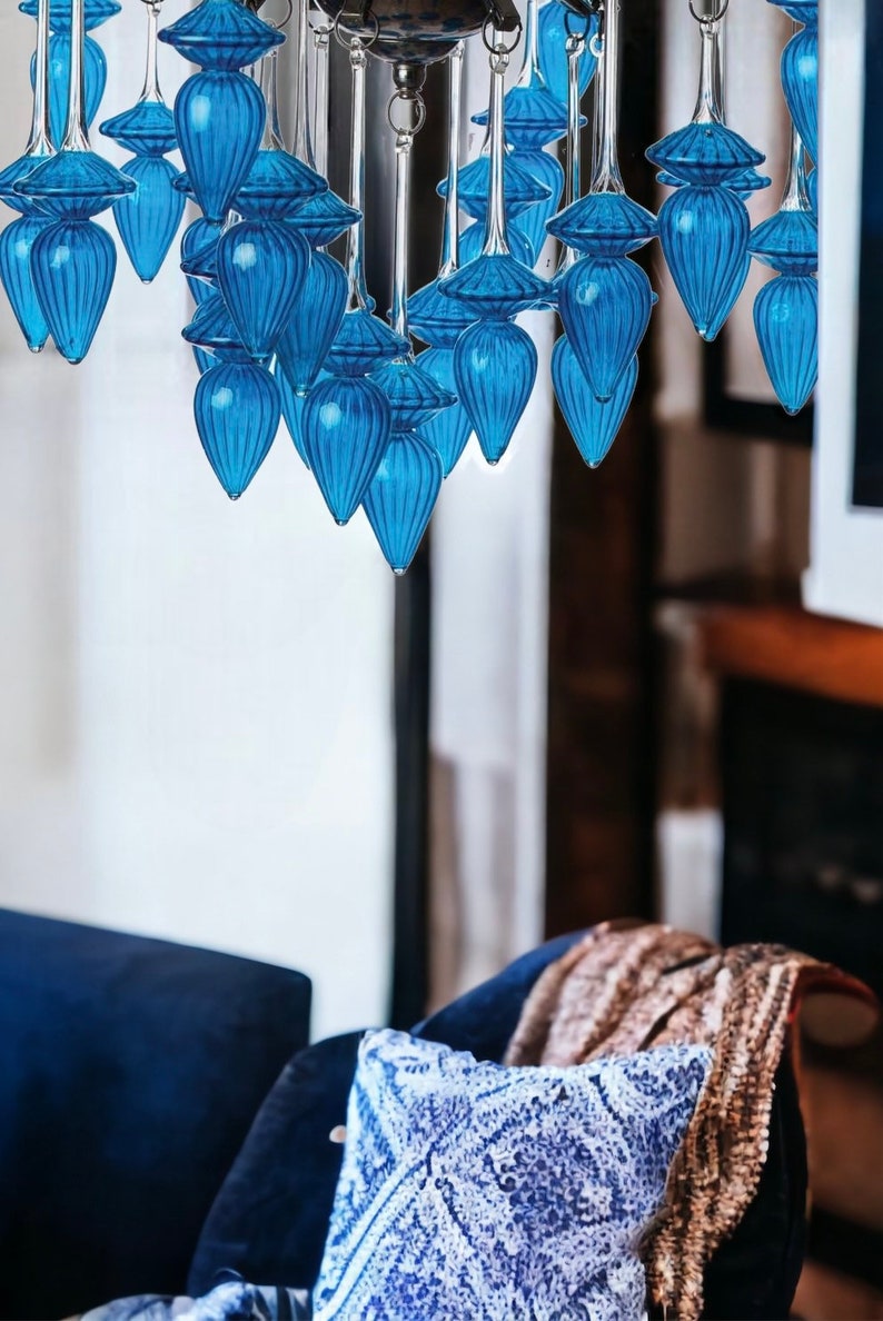 May include: A modern chandelier with multiple blue glass teardrop-shaped pendants. The pendants are suspended from a silver-toned metal frame. The chandelier hangs above a blue velvet chair with a patterned pillow and a brown blanket.