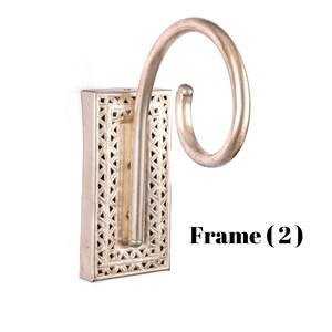 May include: Silver-colored wall sconce with a decorative rectangular base featuring a geometric pattern. The sconce has a curved, scroll-like arm extending from the base. The text "Frame (2)" is visible.