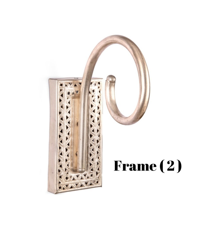 May include: Silver-colored wall sconce with a decorative rectangular base featuring a geometric pattern. The sconce has a curved, scroll-like arm extending from the base. The text "Frame (2)" is visible.