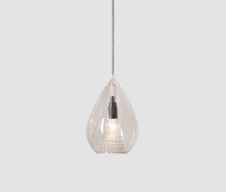 May include: A clear, teardrop-shaped pendant light fixture. The glass shade has a textured, tiered base and a silver-colored interior. A thin, silver cord suspends the light from above, creating a modern and elegant design.