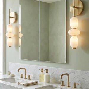 May include: A double vanity bathroom setup featuring a white marble countertop with two integrated sinks, brass faucets, and two matching soap dispensers. Above the vanity, a rectangular mirror is flanked by two wall-mounted brass sconces with three stacked frosted glass globes each.