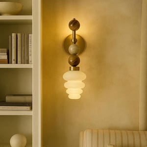 May include: A wall-mounted light fixture with a brass finish and a stacked, spherical design. It features wooden and marble-effect spheres, topped with a frosted glass shade. The sconce is mounted on a light-coloured wall, with a bookshelf and chair in the background.