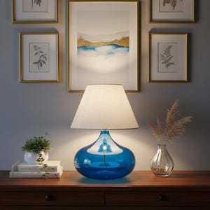 May include: A blue glass table lamp with a white lampshade sits on a wooden table. The lamp has a teardrop-shaped base. A small white pot with a succulent plant and a vase with dried flowers are also on the table.