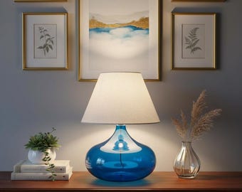 May include: A blue glass table lamp with a white lampshade sits on a wooden table. The lamp has a teardrop-shaped base. A small white pot with a succulent plant and a vase with dried flowers are also on the table.