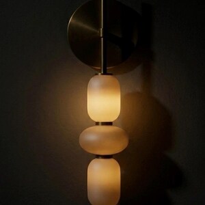 May include: A wall-mounted light fixture featuring a circular brass backplate and a vertical stem. Three stacked, frosted glass shades in rounded, pill-like shapes are illuminated from within, emitting a soft, warm glow against the dark wall.
