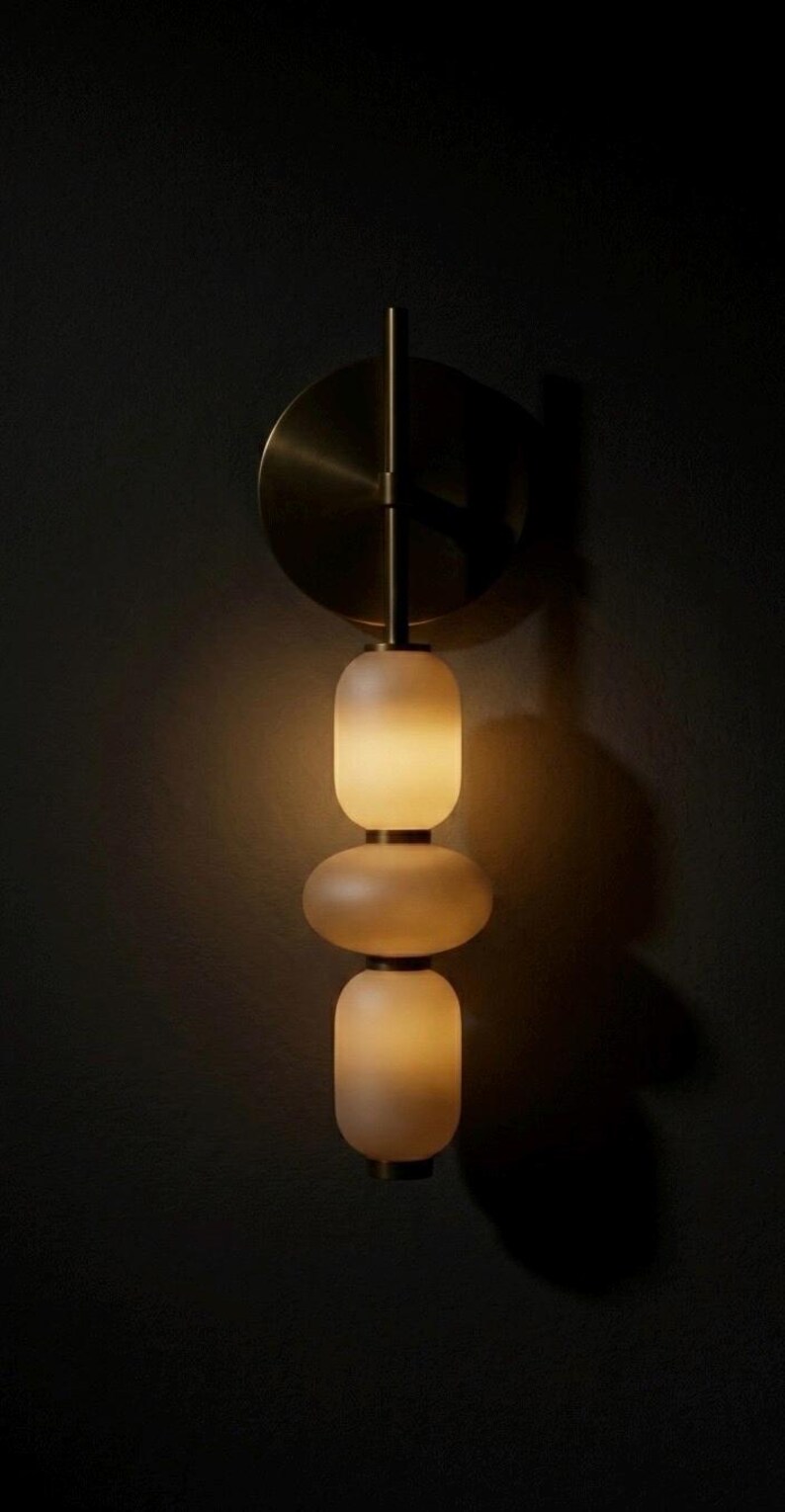 May include: A wall-mounted light fixture featuring a circular brass backplate and a vertical stem. Three stacked, frosted glass shades in rounded, pill-like shapes are illuminated from within, emitting a soft, warm glow against the dark wall.