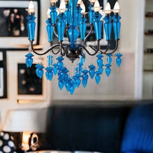 May include: A vibrant blue chandelier with multiple light bulbs. The chandelier has a decorative design with blue glass elements and a silver metal frame. The fixture hangs in a room with framed artwork.