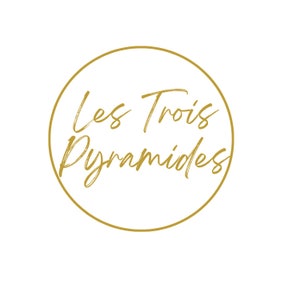 以下が含まれることがあります： 金色のロゴで、筆記体の「Les Trois Pyramides」という文字が金色の円で囲まれています。背景は白です。