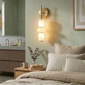May include: A wall-mounted light fixture featuring a circular brass backplate and a vertical rod holding three stacked, rounded, frosted white glass shades that emit a warm, soft glow against the wall surface.