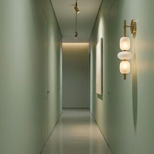 May include: A wall-mounted light fixture featuring a vertical brass rod with two stacked, rounded, frosted white glass globes. The fixture is attached to a sage green wall in a narrow hallway with recessed ceiling spotlights.