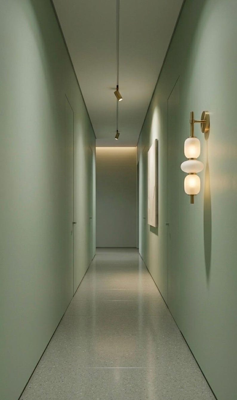 May include: A wall-mounted light fixture featuring a vertical brass rod with two stacked, rounded, frosted white glass globes. The fixture is attached to a sage green wall in a narrow hallway with recessed ceiling spotlights.