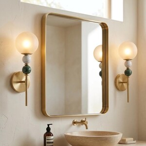 May include: Bathroom wall sconces with a gold finish, featuring a frosted glass globe, and decorative marble and green stone accents. A gold-framed mirror hangs above a stone sink. The sconces are approximately 12 inches tall.